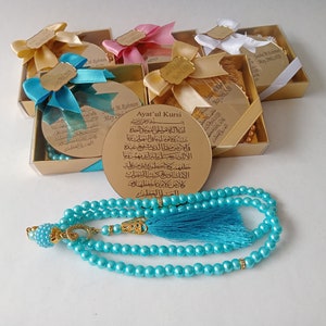 Op de afbeelding: Een set kleine gouden doosjes met blauwe en witte linten, elk met een ronde, gouden plaquette met Arabische tekst. De doosjes zijn in een halve cirkel rond een blauwe gebedskralenketting met een gouden kwast gerangschikt.