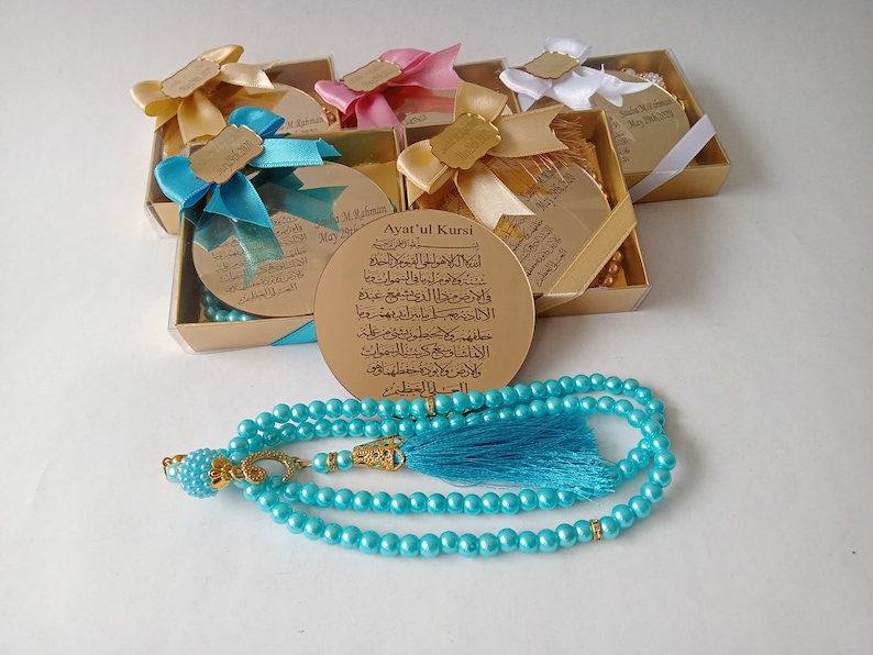 Op de afbeelding: Een set kleine gouden doosjes met blauwe en witte linten, elk met een ronde, gouden plaquette met Arabische tekst. De doosjes zijn in een halve cirkel rond een blauwe gebedskralenketting met een gouden kwast gerangschikt.