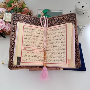 May include: An open Quran rests on a dark brown, intricately carved wooden book stand. A string of light pink prayer beads with a matching pink tassel hangs vertically over the centre of the pages, which feature Arabic text in black and red ink.