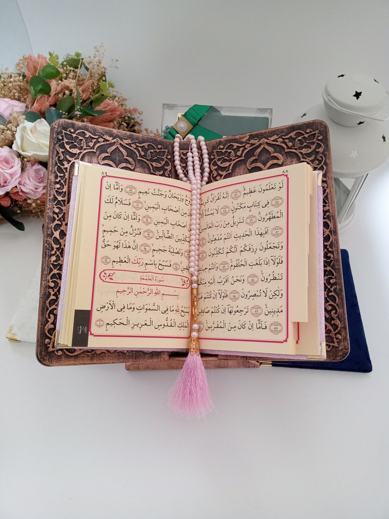May include: An open Quran rests on a dark brown, intricately carved wooden book stand. A string of light pink prayer beads with a matching pink tassel hangs vertically over the centre of the pages, which feature Arabic text in black and red ink.