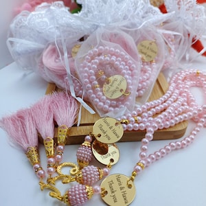May include: Pink beaded prayer beads with gold tassels and a gold charm with the text "Haris & Hawa Thank you".
