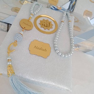 May include: A light grey velvet-covered book featuring a string of light grey pearls, a gold-coloured circular charm with the Arabic word for Allah, and a gold-coloured oval tag engraved with the name Nadiah. A gold letter C charm and a light blue tassel.