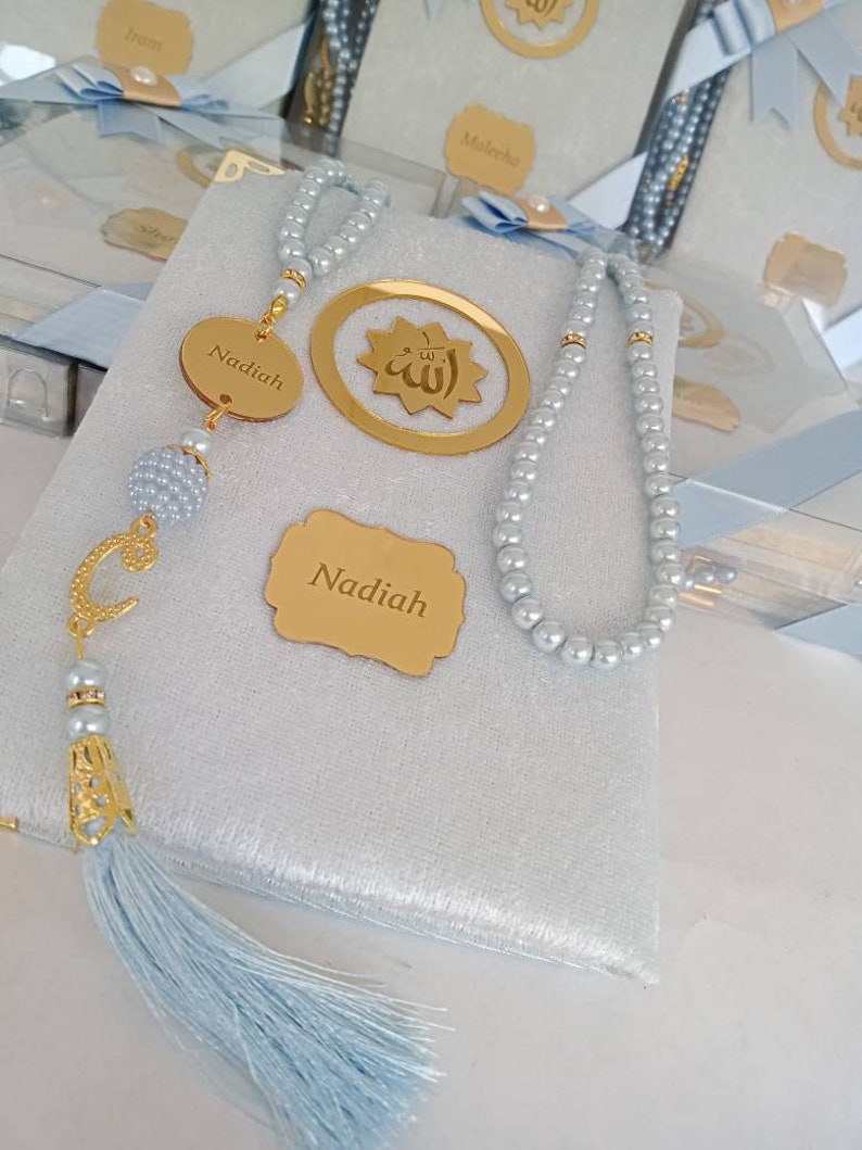 May include: A light grey velvet-covered book featuring a string of light grey pearls, a gold-coloured circular charm with the Arabic word for Allah, and a gold-coloured oval tag engraved with the name Nadiah. A gold letter C charm and a light blue tassel.