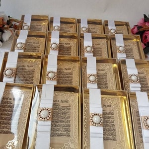 May include: A collection of gold-colored gift boxes with white ribbon and a pearl-like embellishment. Each box contains a printed card with Arabic text, "Ayatul Kursi".