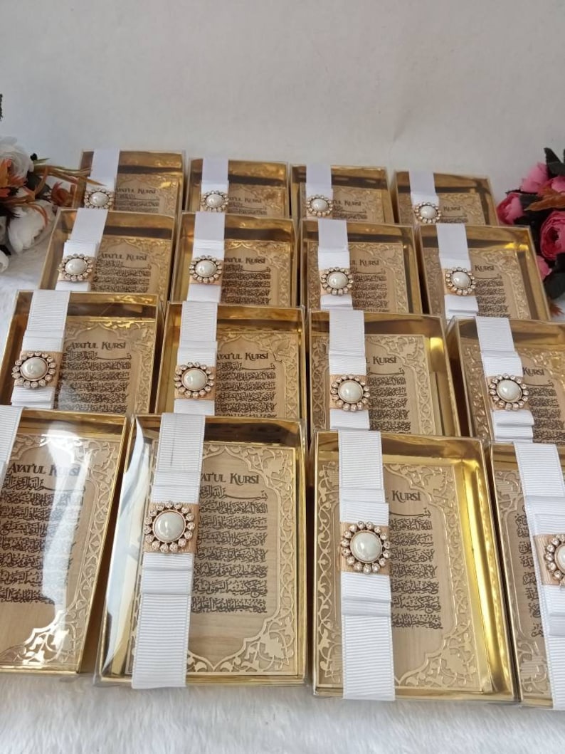 May include: A collection of gold-colored gift boxes with white ribbon and a pearl-like embellishment. Each box contains a printed card with Arabic text, "Ayatul Kursi".