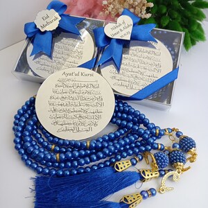 Op de afbeelding: Een set blauwe gebedskralen met gouden accenten en kwastjes. De kralen bevinden zich in een doorzichtige doos met een zilveren plaat die is beschreven met Arabische tekst. De plaat is omwikkeld met een blauw lint. De tekst op de plaat is "Ayatul Kursi".