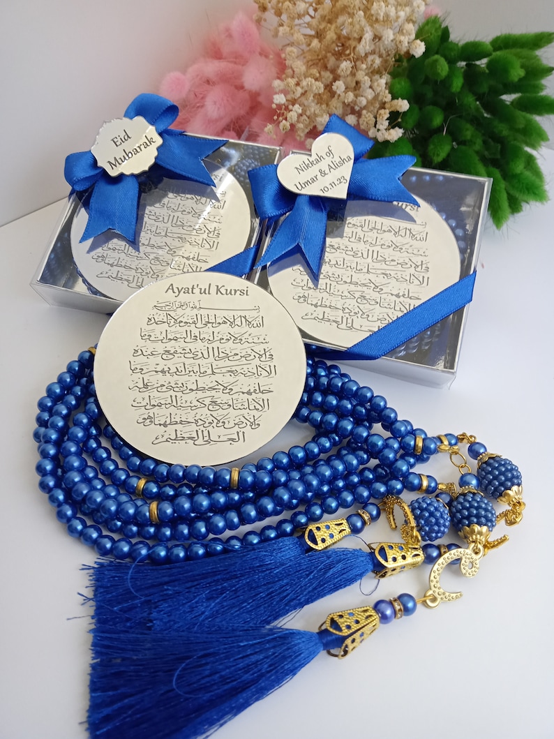 Op de afbeelding: Een set blauwe gebedskralen met gouden accenten en kwastjes. De kralen bevinden zich in een doorzichtige doos met een zilveren plaat die is beschreven met Arabische tekst. De plaat is omwikkeld met een blauw lint. De tekst op de plaat is "Ayatul Kursi".