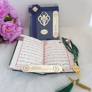 Puede incluir: Una caja de regalo azul oscuro con un lazo marino y el nombre Mohammed, un Cor&aacute;n abierto con texto en &aacute;rabe, un marcap&aacute;ginas de metal dorado con caligraf&iacute;a &aacute;rabe y un rosario de cuentas verdes con una etiqueta circular dorada con el nombre Nessrine.