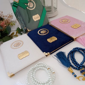 May include: A collection of four velvet-covered books in white, navy blue, pink, and green, each featuring gold-toned corner protectors, a circular emblem with Arabic calligraphy, and a personalised name plaque. Two sets of prayer beads are included.