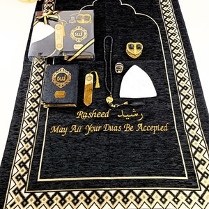 May include: A black prayer rug with gold geometric borders and the embroidered text "Rasheed" and "May All Your Duas Be Accepted." Included are a black Quran, a gold bookmark, black prayer beads, a white kufi cap, a gold ring, and a heart-shaped box.