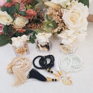 May include: Three clear boxes with white ribbon and gold heart-shaped charms with personalized text. Each box contains a string of white, black, or cream-colored beads with a tassel.