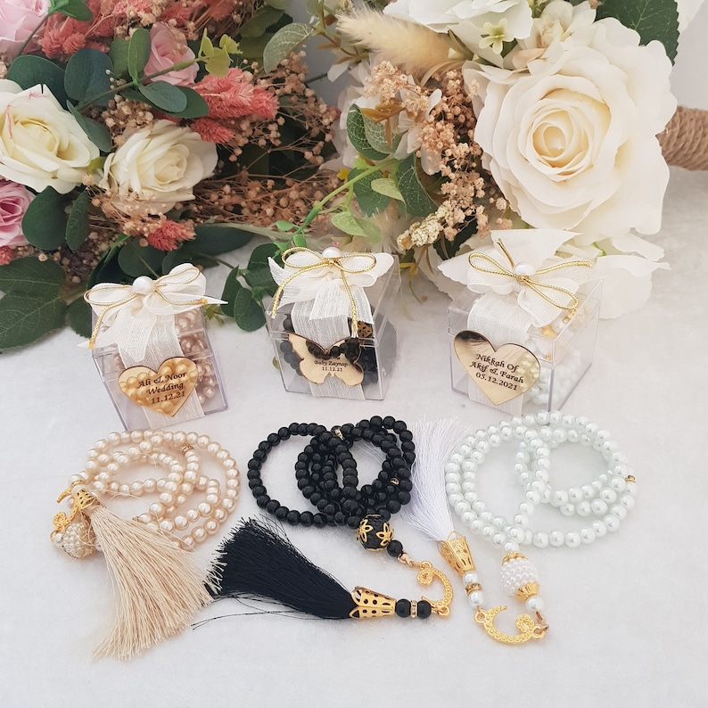 May include: Three clear boxes with white ribbon and gold heart-shaped charms with personalized text. Each box contains a string of white, black, or cream-colored beads with a tassel.