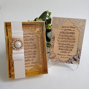 May include: Two wooden plaques with gold-colored trim and Arabic calligraphy. The plaques are engraved with the text "Ayatul Kursi" and "Bismillah ar-Rahman ar-Rahim". The plaques are on a white background.