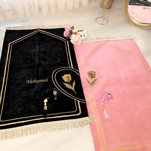 May include: Two prayer rugs with gold trim and tassels. One rug is black with the name "Mohamed" embroidered on it and the other is pink.