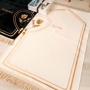 May include: Two white prayer rugs with gold trim and tassels. The rug on the left has the name "Mohamed" embroidered on it. The rug on the right has the name "Ayesha" embroidered on it.