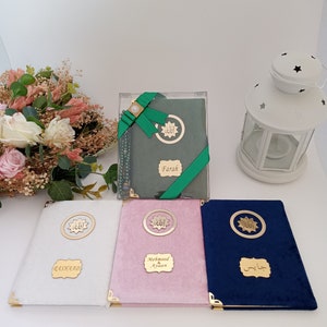 May include: Four rectangular books with velvet covers in white, pink, dark blue, and sage green. Each book features gold-toned metal corner protectors, a circular emblem with Arabic calligraphy, and a rectangular nameplate with custom text.