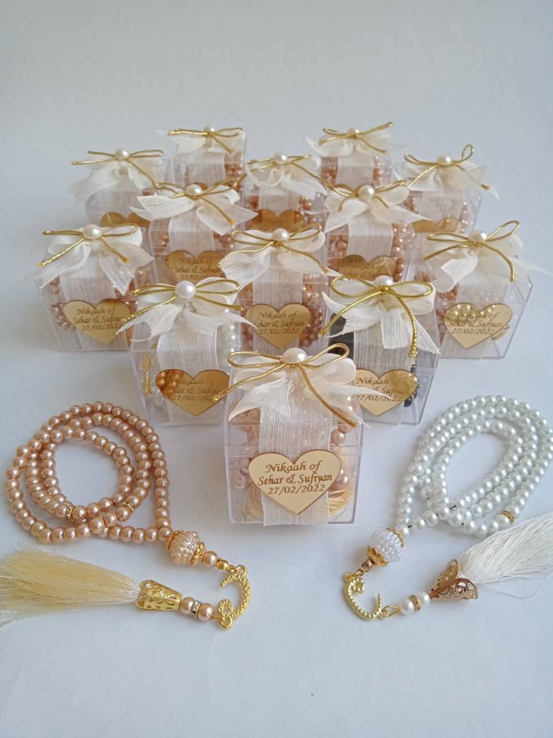 May include: A set of 12 clear plastic boxes with gold trim, each containing a white pearl necklace and a small gold heart with the text "Nikah of Sehar & Sufiyan 27/02/2022". Two additional necklaces are shown, one with white pearls and one with brown pearls.