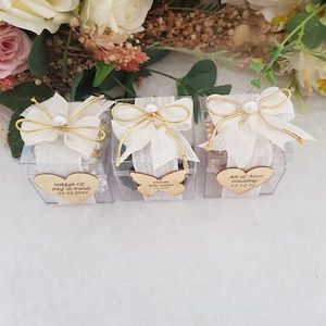 May include: Three clear plastic boxes with white fabric and gold ribbon bows. Each box has a wooden heart or butterfly with text. The first box says "Nikah of Arif & Farah 05.12.2021". The second box says "Welcome Baby Zoya 11.12.21". The third box says "Ali & Noor Wedding 11.12.21".