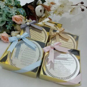 May include: Four silver circular plaques with Arabic calligraphy and a decorative border of rhinestones are displayed in gold boxes with light blue, pink, and white ribbons. The plaques are wedding favors with the names "TAIM & OMAR" and the date "12.09.2021" on one of the plaques.