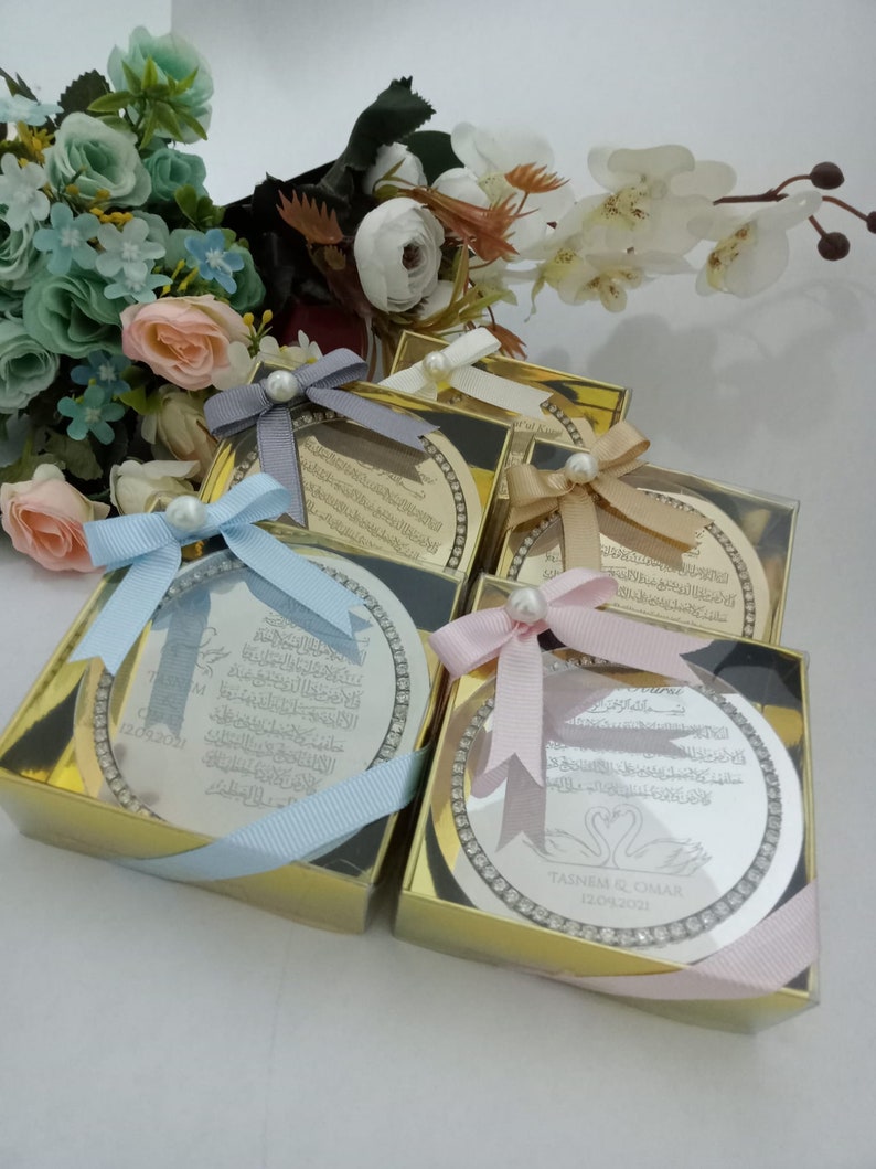 May include: Four silver circular plaques with Arabic calligraphy and a decorative border of rhinestones are displayed in gold boxes with light blue, pink, and white ribbons. The plaques are wedding favors with the names "TAIM & OMAR" and the date "12.09.2021" on one of the plaques.