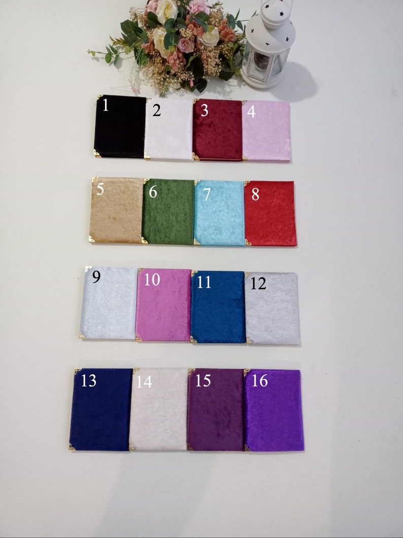May include: Sixteen rectangular velvet-covered notebooks arranged in four rows, each featuring gold-tone metal corner protectors. The colours include black, white, burgundy, light pink, tan, green, light blue, red, silver, magenta, navy, grey, blue, cream, plum, and purple.