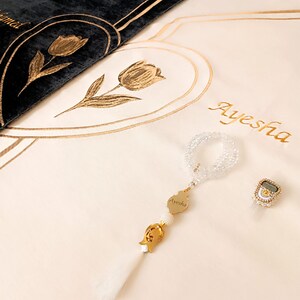 May include: A white prayer rug with gold embroidery of a tulip and the name Ayesha. A white prayer bead necklace with a gold charm that says Ayesha is on the rug. A gold ring with a digital prayer counter is also on the rug.