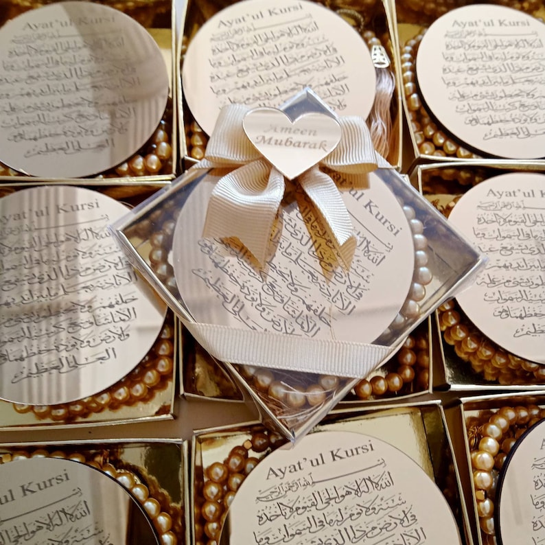 Op de afbeelding: Een set kleine, ronde, gouden doosjes met een doorzichtig deksel. Elk doosje bevat een kleine, gouden kralenketting en een ronde, gouden spiegel met Arabische tekst. De spiegel heeft een witte achtergrond en de tekst is zwart. De doosjes zijn versierd met een wit lint en een hartvormig label met de tekst "Ameen Mubarak".