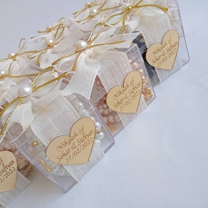 May include: A row of clear plastic boxes decorated with white fabric and gold ribbon. Each box contains a small pearl necklace, a gold charm, and a heart-shaped tag with the text "Nikaah of Sehar & Sufiyan 27/02/2022".
