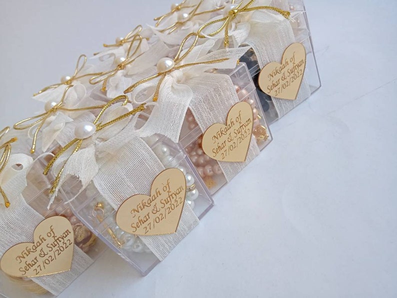 May include: A row of clear plastic boxes decorated with white fabric and gold ribbon. Each box contains a small pearl necklace, a gold charm, and a heart-shaped tag with the text "Nikaah of Sehar & Sufiyan 27/02/2022".