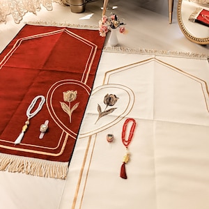 May include: Two prayer rugs with gold trim and floral designs. One rug is red and the other is white. Both rugs have a set of prayer beads and a prayer counter.