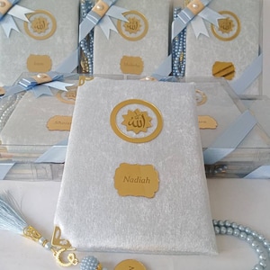 May include: A set of light blue velvet-covered books in clear plastic boxes, each featuring a gold circular emblem with Arabic calligraphy, a gold nameplate, and a light blue ribbon. A matching light blue pearl prayer bead strand with a gold name tag.