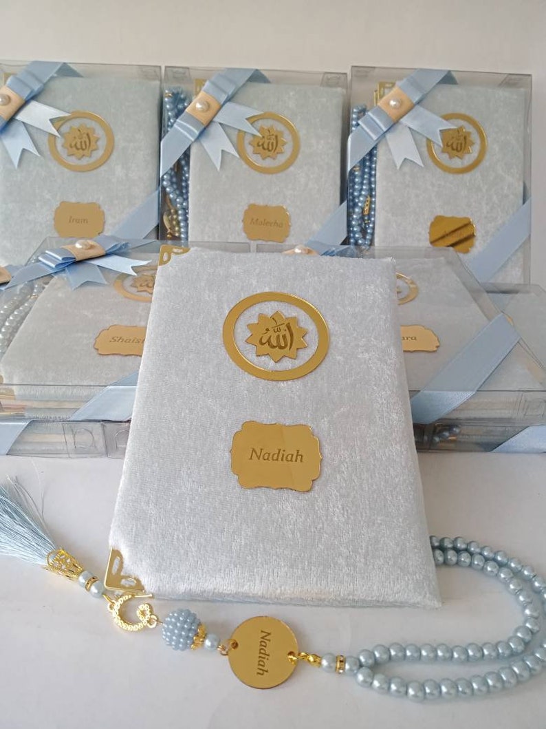 May include: A set of light blue velvet-covered books in clear plastic boxes, each featuring a gold circular emblem with Arabic calligraphy, a gold nameplate, and a light blue ribbon. A matching light blue pearl prayer bead strand with a gold name tag.