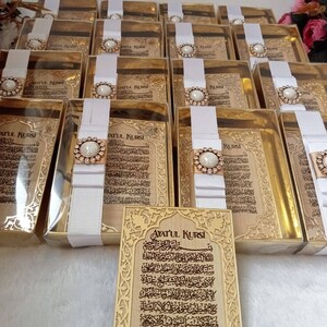 May include: A set of gold-colored boxes with white ribbon and a pearl-like embellishment. Each box contains a small, framed piece of paper with Arabic calligraphy. The text on the paper reads "Ayatul Kursi".