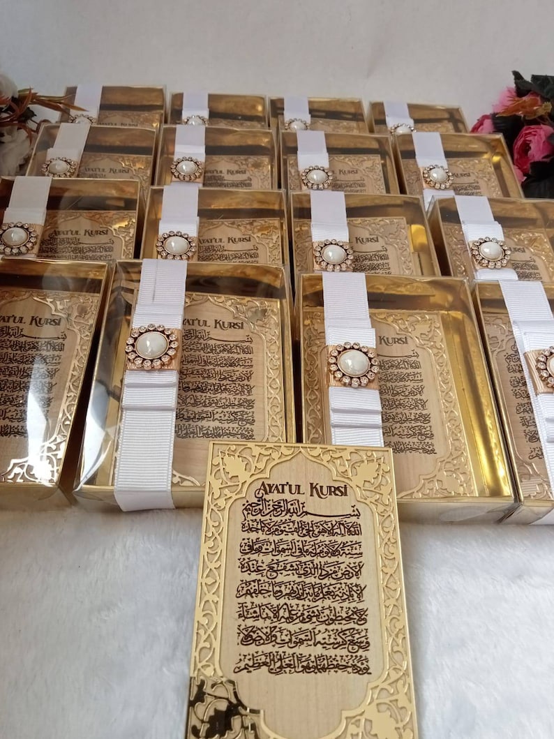 May include: A set of gold-colored boxes with white ribbon and a pearl-like embellishment. Each box contains a small, framed piece of paper with Arabic calligraphy. The text on the paper reads "Ayatul Kursi".
