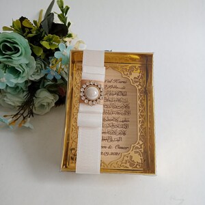 May include: A gold-colored frame with a white ribbon and a pearl-like embellishment. The frame contains a printed inscription in Arabic script with the text "t'ul Kursi" and the date "2.09.2021".