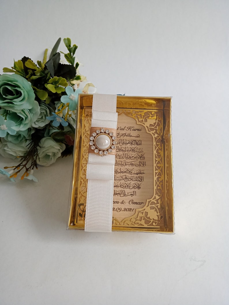 May include: A gold-colored frame with a white ribbon and a pearl-like embellishment. The frame contains a printed inscription in Arabic script with the text "t'ul Kursi" and the date "2.09.2021".