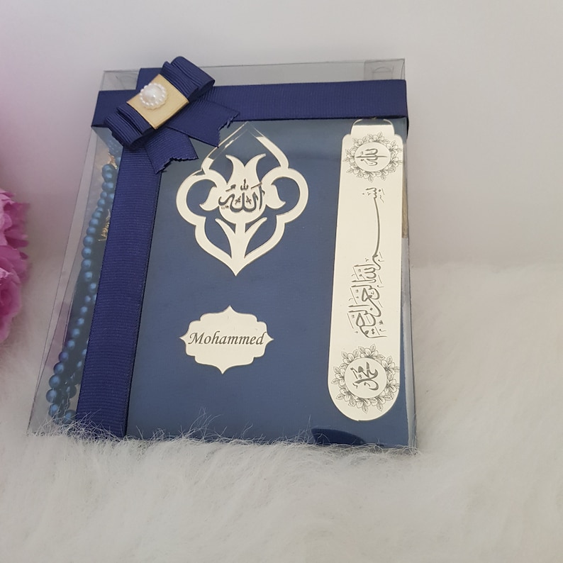 Puede incluir: Un set de regalo azul oscuro en una caja de pl&aacute;stico transparente que incluye un libro de oraciones con caligraf&iacute;a &aacute;rabe dorada y el nombre Mohammed, un marcap&aacute;ginas de metal dorado a juego y cuentas de oraci&oacute;n azules, con un lazo azul marino.