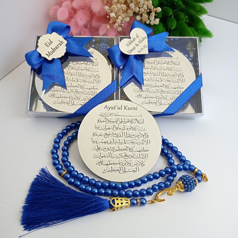 Op de afbeelding: Een blauwe en gouden gebedsparelset met een zilveren ronde plaquette gegraveerd met Arabische schrift en de woorden "Ayat'ul Kursi". De plaquette bevindt zich in een doorzichtige doos met een blauw lint en een hartvormig label met de tekst "Nikkah of Umar & Alisha 10.11.23".