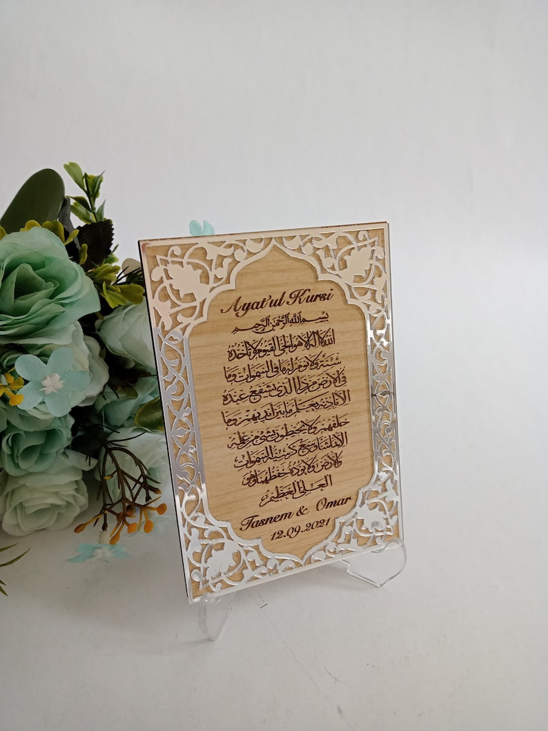 May include: A wooden plaque with a silver frame and intricate floral designs. The plaque features the Arabic text of Ayatul Kursi, a verse from the Quran, and the names "Fasnem & Omar" with the date "12.09.2021".