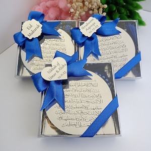 Op de afbeelding: Drie kleine, doorzichtige doosjes met blauwe strikken en zilveren ronde plaquettes erin. De plaquettes hebben Arabische kalligrafie en tekst die "Eid Mubarak", "Nikkah of Umar & Alisha 10.11.23" en "Ibraheem Aqeeqa 10.12.23" luidt.