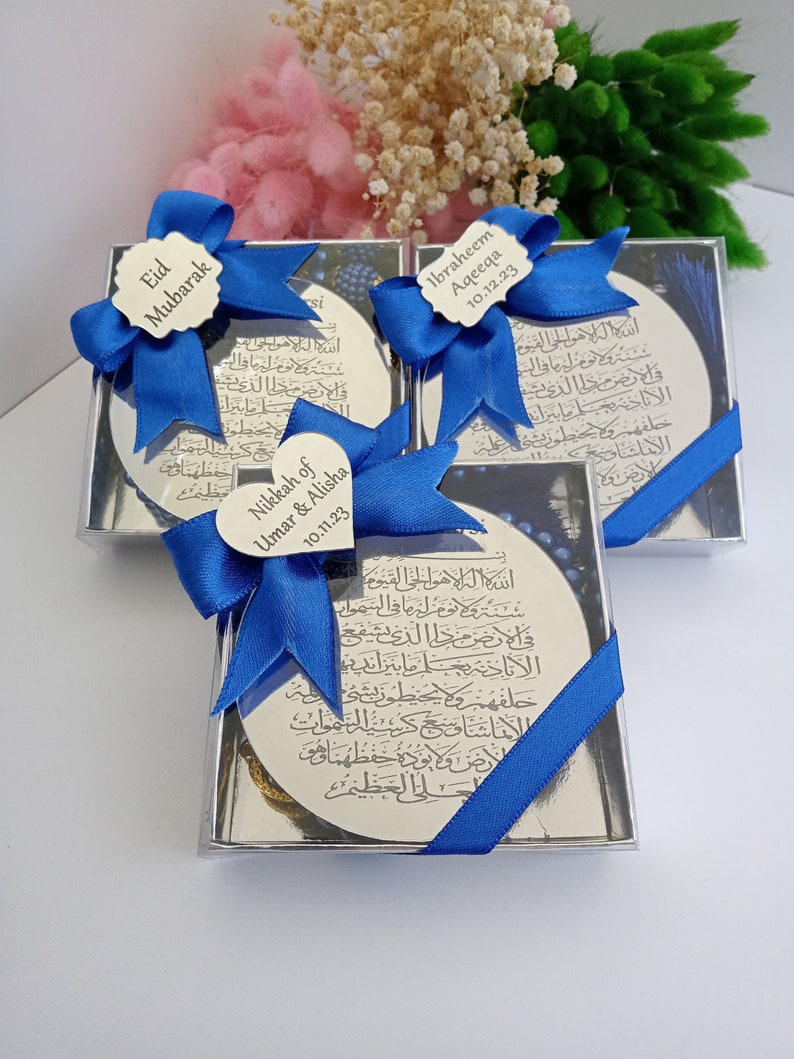 Op de afbeelding: Drie kleine, doorzichtige doosjes met blauwe strikken en zilveren ronde plaquettes erin. De plaquettes hebben Arabische kalligrafie en tekst die "Eid Mubarak", "Nikkah of Umar & Alisha 10.11.23" en "Ibraheem Aqeeqa 10.12.23" luidt.