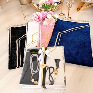 May include: A gift set for a wedding or special occasion. The set includes three prayer rugs in black, white, and blue with gold trim, two prayer beads, and a white gift box with a gold ribbon and a heart-shaped tag that says "Ayla & Muhammed".