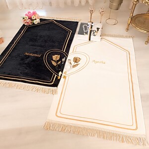 May include: Two prayer rugs with gold trim and fringe. The rug on the left is black with the name "Mohamed" embroidered on it. The rug on the right is white with the name "Ayesha" embroidered on it.