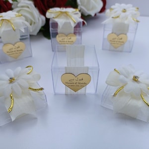 Puede incluir: Cajas de regalo blancas con cinta dorada y una flor blanca en la parte superior. Cada caja tiene una etiqueta dorada en forma de corazón con el texto "Qubool Hai" y los nombres "Hussain & Mamha" y la fecha "19 de noviembre de 2021".