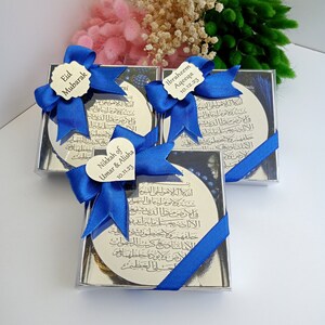 Op de afbeelding: Drie zilveren doosjes met blauwe strikken en gepersonaliseerde labels. De doosjes bevatten kleine zilveren plaquettes met Arabische kalligrafie. De labels luiden "Eid Mubarak", "Nikkah of Umar & Alisha 10.11.23" en "Ibraheem Aqeeqa 10.12.23".