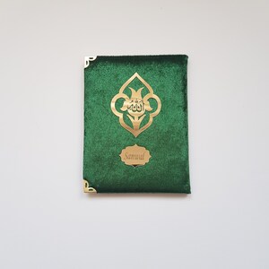 Puede incluir: Una cubierta de libro rectangular de terciopelo verde con protectores de esquina de metal dorado. El frente muestra un emblema central dorado con caligraf&iacute;a &aacute;rabe y una placa dorada m&aacute;s peque&ntilde;a con el nombre grabado Sanwal.