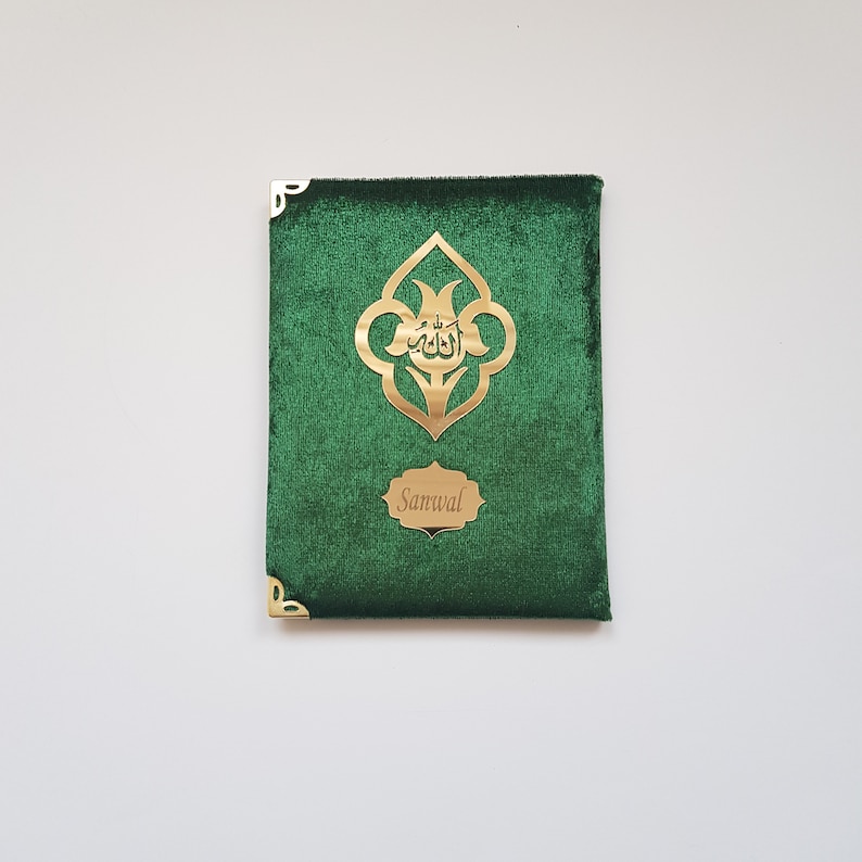Puede incluir: Una cubierta de libro rectangular de terciopelo verde con protectores de esquina de metal dorado. El frente muestra un emblema central dorado con caligraf&iacute;a &aacute;rabe y una placa dorada m&aacute;s peque&ntilde;a con el nombre grabado Sanwal.