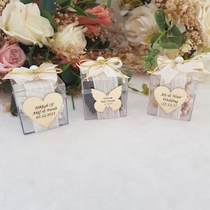 May include: Three clear plastic boxes with white ribbon and pearl accents. Each box contains a wooden heart or butterfly with personalized text. The text on the boxes reads "Nikkah of Akif & Farah 05.12.2021", "Welcome Baby Zaynap 11.12.21", and "Ali & Noor Wedding 11.12.21".