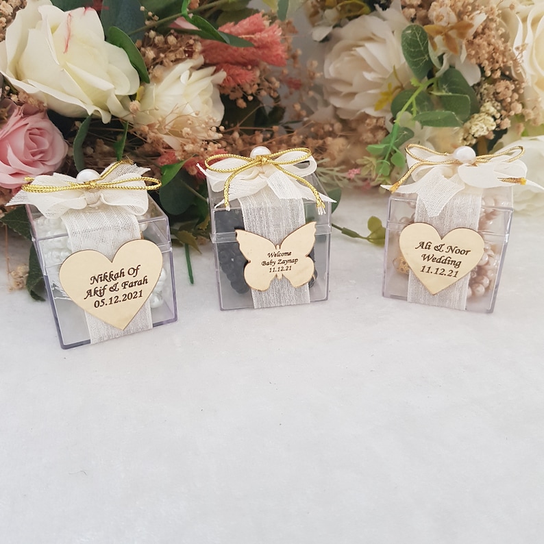 May include: Three clear plastic boxes with white ribbon and pearl accents. Each box contains a wooden heart or butterfly with personalized text. The text on the boxes reads "Nikkah of Akif & Farah 05.12.2021", "Welcome Baby Zaynap 11.12.21", and "Ali & Noor Wedding 11.12.21".