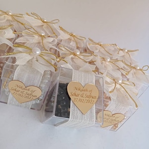 May include: A group of small, clear boxes with white fabric and gold ribbon bows. Each box contains black beads and a gold heart-shaped tag with the text "Nikaah of Sehar & Sufiyan 27/02/2022".
